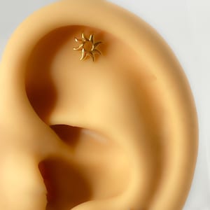 Könnte beinhalten: Nahaufnahme eines Ohrs mit zwei sonnenförmigen Ohrsteckern. Einer ist goldfarben, der andere silberfarben. Die Ohrringe sind klein und detailliert, mit Strahlen, die von einem zentralen Kreis ausgehen.