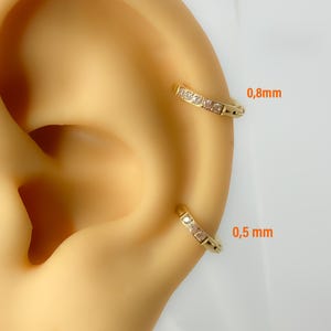 Peut inclure: Deux boucles d'oreilles créoles dorées avec de petits pierres précieuses transparentes sont présentées sur une oreille de couleur claire. L'oreille supérieure est étiquetée "0,8 mm" et l'oreille inférieure est étiquetée "0,5 mm".