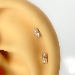 Könnte beinhalten: Zwei goldfarbene Ohrstecker, jeweils mit einer kleinen Perle und einem klaren, funkelnden Edelstein. Die Ohrringe werden an einem Ohr gezeigt und zeigen ihr zartes Design und ihr elegantes Aussehen.