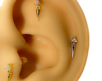 Könnte beinhalten: Gold- und silberfarbene Ohrringe mit dreieckigem Oberteil und einem herabhängenden, spitz zulaufenden Boden. Die Ohrringe sind an einem Ohr abgebildet, wobei drei verschiedene Ohrringe sichtbar sind. Die Ohrringe haben ein minimalistisches Design.