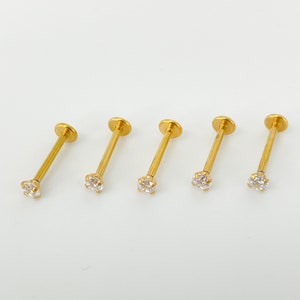 Könnte beinhalten: Fünf goldfarbene Lippenpiercing-Stecker mit einem kleinen, klaren Edelstein am Ende jedes Stifts. Die Stecker sind horizontal vor einem weißen Hintergrund angeordnet, um ihr Design und ihre Farbe hervorzuheben.