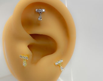 Könnte beinhalten: Ohrringe mit einem Stangendesign und einem herabhängenden tropfenförmigen Edelstein. Die Ohrringe werden in Silber- und Goldtönen mit klaren Edelsteinen gezeigt. Die Ohrringe werden auf einem beigefarbenen Ohrmodell präsentiert.