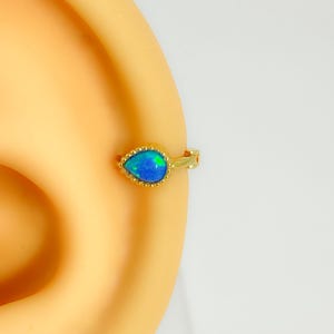 Könnte beinhalten: Goldfarbener Ohrring mit einem tropfenförmigen Opalstein in Blau und Grün. Der Ohrring ist an einem Ohr befestigt. Der Opalstein hat eine strukturierte Oberfläche und ist von einem Perlenrand umgeben.
