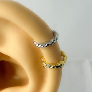 Puede incluir: Primer plano de dos pequeños pendientes de aro. Uno es plateado, el otro dorado. Ambos tienen un diseño de cuerda trenzada y pequeñas piedras preciosas transparentes. Los pendientes se llevan en una oreja, con un fondo neutro.