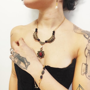 Könnte beinhalten: Ein goldfarbenes Halsketten-, Armband- und Ohrring-Set. Die Halskette hat einen Anhänger mit einem roten Stein und aufwendigen Metallarbeiten. Armband und Halskette haben dunkle Perlen. Die Ohrringe haben lila Perlen und einen weißen Totenkopf-Anhänger. Der Schmuck wird mit einem schwarzen Oberteil getragen.