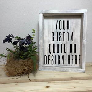 May include: A rustic wooden framed sign with the text "YOUR CUSTOM QUOTE OR DESIGN HERE" in black. The frame has a distressed white finish. A small burlap-wrapped plant with dark purple flowers sits to the left.