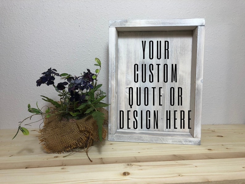 May include: A rustic wooden framed sign with the text "YOUR CUSTOM QUOTE OR DESIGN HERE" in black. The frame has a distressed white finish. A small burlap-wrapped plant with dark purple flowers sits to the left.