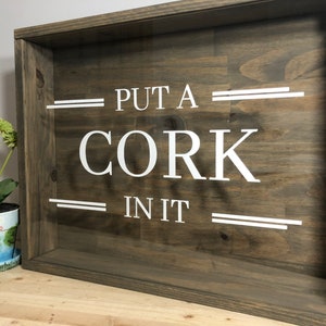 May include: A wooden box with a glass front, featuring the text "PUT A CORK IN IT" in white lettering. The box is stained a dark brown colour and has a rustic appearance.