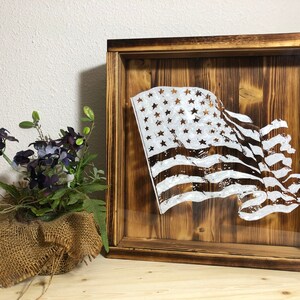 Puede incluir: Una caja de sombra de madera con un diseño de bandera estadounidense blanca desgastada en un panel de vidrio transparente. La caja tiene un marco de madera oscura y está sobre una superficie de madera clara.