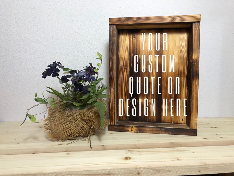 May include: A rustic wooden sign with the text "YOUR CUSTOM QUOTE OR DESIGN HERE" in white. The sign is framed and has a dark, burnt wood finish. A small potted plant with purple flowers and green foliage sits to the left.