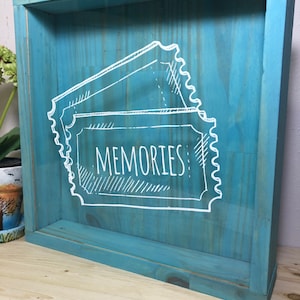 May include: A teal blue wooden shadow box with a glass front. The box has a white drawing of three ticket stubs with the word "MEMORIES" written on the center stub.