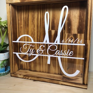 May include: A wooden shadow box with a clear front. The box features a large white letter "M" with the names "Ty & Cassie" and the date "6/14/14" in white script. The wood has a warm, brown tone.