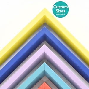 May include: A collection of colorful picture frames in various shades, including yellow, blue, lavender, teal, and red. The frames are arranged in a layered, angled pattern, with the text "Custom Sizes AVAILABLE" in a teal circle.
