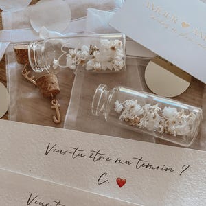 May include: Two small glass bottles with cork stoppers filled with dried white flowers. Each bottle has a gold charm attached to the cork. The bottles are on a white surface with a handwritten note that says "Veux-tu &ecirc;tre ma t&eacute;moin? C." and "Veux-tu &ecirc;tre mon t&eacute;moin? J."