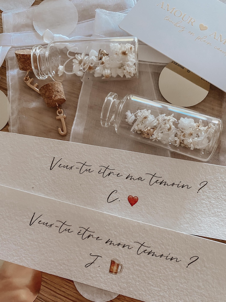 May include: Two small glass bottles with cork stoppers filled with dried white flowers. Each bottle has a gold charm attached to the cork. The bottles are on a white surface with a handwritten note that says "Veux-tu &ecirc;tre ma t&eacute;moin? C." and "Veux-tu &ecirc;tre mon t&eacute;moin? J."