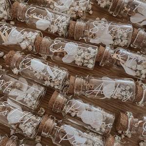 May include: A collection of small glass bottles with cork tops filled with dried flowers and a handwritten name on each bottle. The names include: Joyce, Camille, Millie, Eugene, Camille,  Maman, Estelle, Papa,  Pauline, Audrey, Solene,  Raph, and Maman.