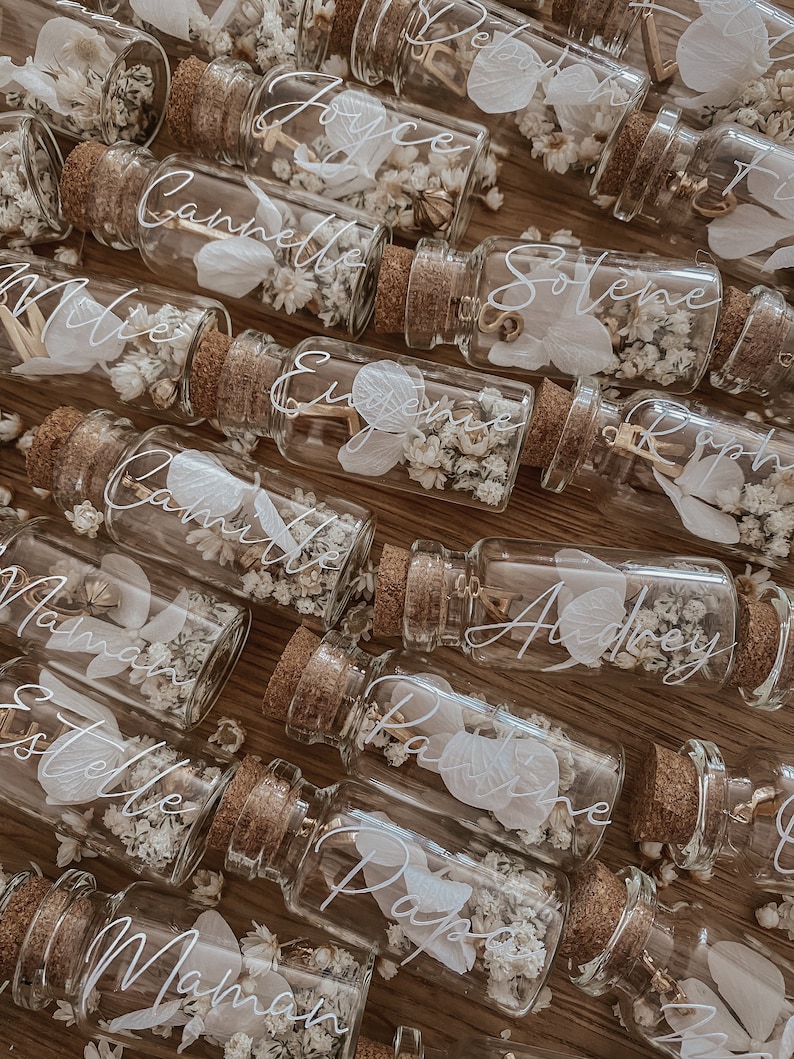May include: A collection of small glass bottles with cork tops filled with dried flowers and a handwritten name on each bottle. The names include: Joyce, Camille, Millie, Eugene, Camille,  Maman, Estelle, Papa,  Pauline, Audrey, Solene,  Raph, and Maman.