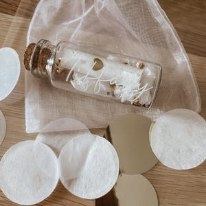 May include: A small glass bottle with a cork stopper filled with white confetti and a gold heart charm. The bottle is labelled with the name "Tiffany" in white script. The bottle is sitting on a wooden surface with white and gold confetti.