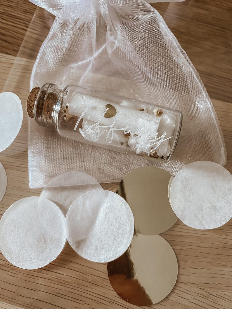 May include: A small glass bottle with a cork stopper filled with white confetti and a gold heart charm. The bottle is labelled with the name "Tiffany" in white script. The bottle is sitting on a wooden surface with white and gold confetti.