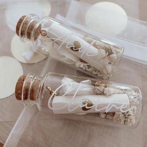 May include: Two glass bottles with cork stoppers filled with dried white flowers and a rolled up piece of paper with the names "Anne" and "Claire" written on them.
