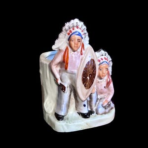 May include: A ceramic planter shaped like a tree stump featuring two figures wearing feathered headdresses. One figure stands holding a round shield, while the other kneels beside them. The piece is painted in shades of tan, brown, and white.