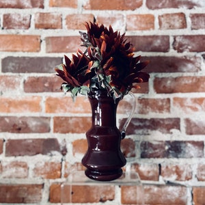 May include: A dark burgundy ceramic vase with a flared base and a handle, holding a bouquet of dried red sunflowers against a brick wall background.