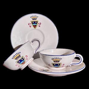 May include: White teacups and saucers with a blue rim and a crest design. The crest features a crown and shield with red, blue, and yellow accents. The set includes two cups and two saucers.