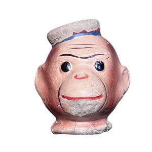 May include: A vintage-style molded monkey head figurine featuring a light brown complexion, black circular eyes, and a red painted mouth. The monkey wears a small gray cap with a thin blue stripe detail. The surface shows signs of wear and texture.