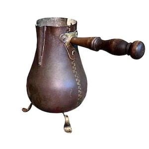 May include: A copper-colored Turkish coffee pot with a wooden handle and three small feet. The pot has a bulbous body and a narrow neck. The handle is attached with brass hardware. The pot is designed for stovetop use.