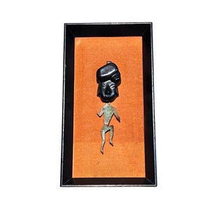 May include: A black carved stone head with a stylized, dark, textured metal body attached, displayed inside a rectangular black frame with an orange backing. The figure has a smooth, rounded head and a thin, angular, metallic torso and limbs.