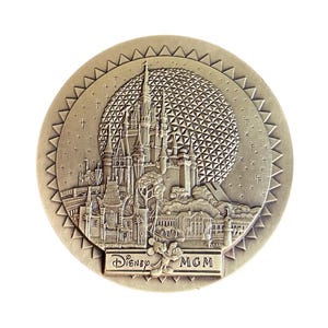 May include: A round, metallic coin with a detailed relief of a castle, a geometric sphere, and the text "Disney MGM". The coin has a textured edge and a light bronze color, with a white background.