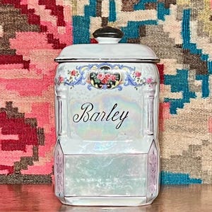 May include: A white ceramic jar with a lid, featuring the word "Barley" in elegant script. The jar is adorned with floral and scroll designs in pink, blue, and gold, with a black knob on top. The jar has a pearlescent finish.