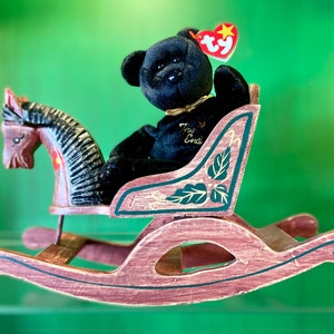 May include: A black beanie baby bear sitting on a wooden rocking horse. The horse is painted brown with green and gold accents. The bear has a red heart-shaped tag that says "Ty" on it. The bear is wearing a gold ribbon around its neck and has the words "The End" embroidered on its chest.