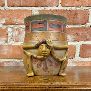 May include: A handcrafted ceramic vase with a unique design. The vase features a tan base with a face and arms, and a geometric pattern in shades of brown, red, and blue. The top rim is green. The vase is displayed on a wooden surface.