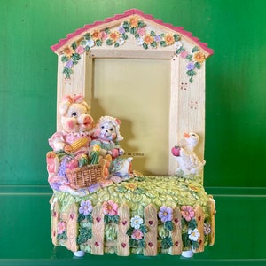 May include: A white picture frame with a pink roof and a floral design. The frame features a pig, a lamb, and a duck figurine. The frame is made of ceramic and has a rustic charm.