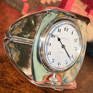 May include: A silver-toned Tiffany & Co. travel clock with a white face and black numerals. The clock has a rectangular shape with curved sides and a hinged cover. The clock face reads "Tiffany & Co." and "8 Days."