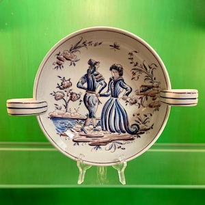 May include: A white ceramic bowl with two handles, featuring a hand-painted scene of two figures in blue and white clothing, standing in a landscape with flowers and trees. The bowl has a white background with brown accents.