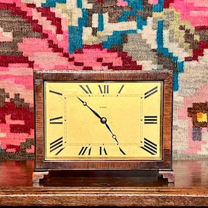 May include: A rectangular wooden clock with a light yellow face and black Roman numerals. The clock has a dark brown wooden frame and stands on a wooden surface. The background features a colorful patterned textile.