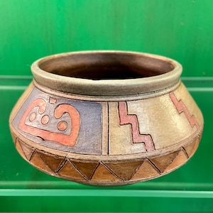 May include: A handcrafted ceramic bowl with a wide, rounded shape. The bowl features geometric designs in shades of brown, blue, and peach. The rim is a muted green. The bowl's design is reminiscent of ancient pottery.