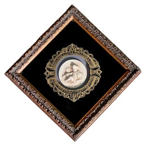 May include: A framed, diamond-shaped artwork featuring a bas-relief of a cherub holding a book and a scroll. The cherub is set against a black background, framed by an ornate, dark brown wooden frame with gold accents.