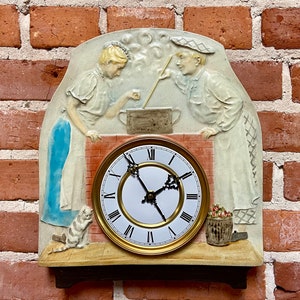 May include: A ceramic wall clock with a white face and black hands. The clock is decorated with a relief sculpture of two figures cooking in a kitchen. The figures are wearing white aprons and are holding cooking utensils. The clock is mounted on a brick wall.