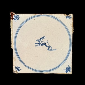 May include: Square ceramic tile with a white background and a blue rabbit illustration inside a blue circle. The tile has a distressed appearance with blue floral corner accents.