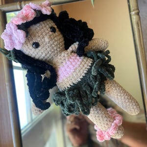Pode incluir: Boneca de croch&ecirc; feita &agrave; m&atilde;o com corpo bege, cabelo preto e uma roupa rosa e verde. A boneca tem uma coroa de flores rosa e detalhes rosa nos tornozelos. A boneca &eacute; refletida em um espelho.
