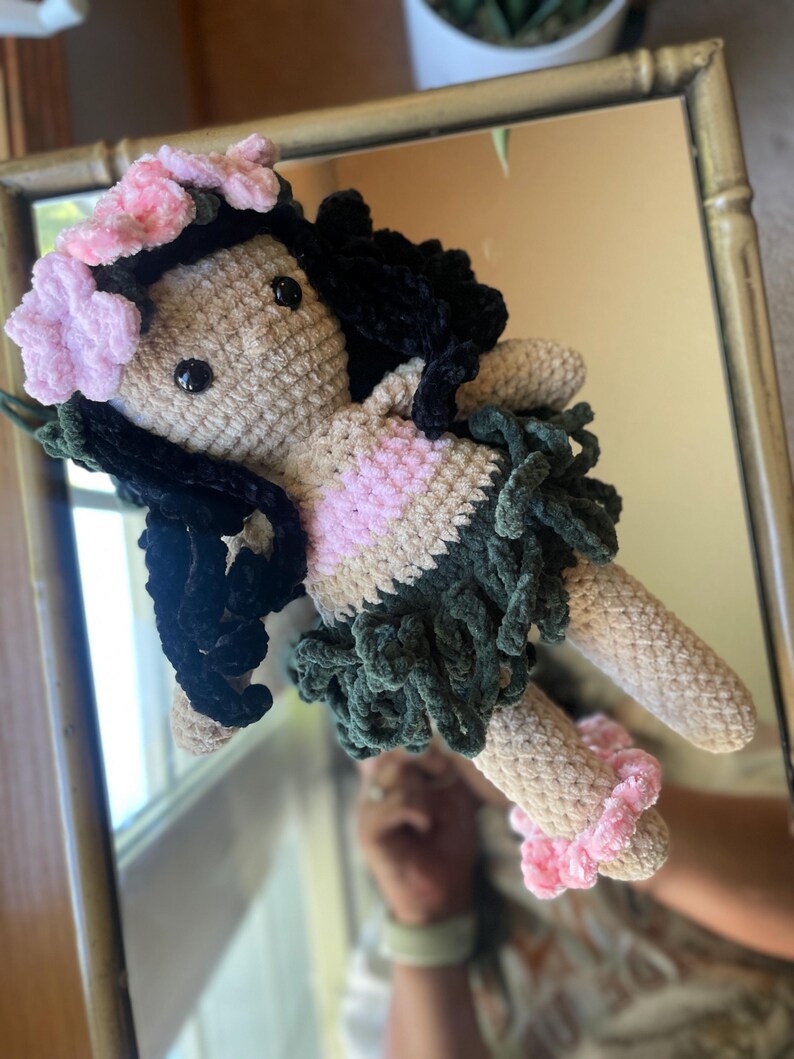 Pode incluir: Boneca de croch&ecirc; feita &agrave; m&atilde;o com corpo bege, cabelo preto e uma roupa rosa e verde. A boneca tem uma coroa de flores rosa e detalhes rosa nos tornozelos. A boneca &eacute; refletida em um espelho.