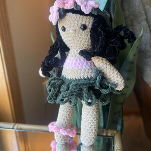Pode incluir: Boneca de croch&ecirc; com corpo bege, cabelo preto e uma roupa rosa e verde. A boneca tem uma coroa de flores rosa e acess&oacute;rios de tornozelo combinando. A boneca est&aacute; em frente a um espelho.
