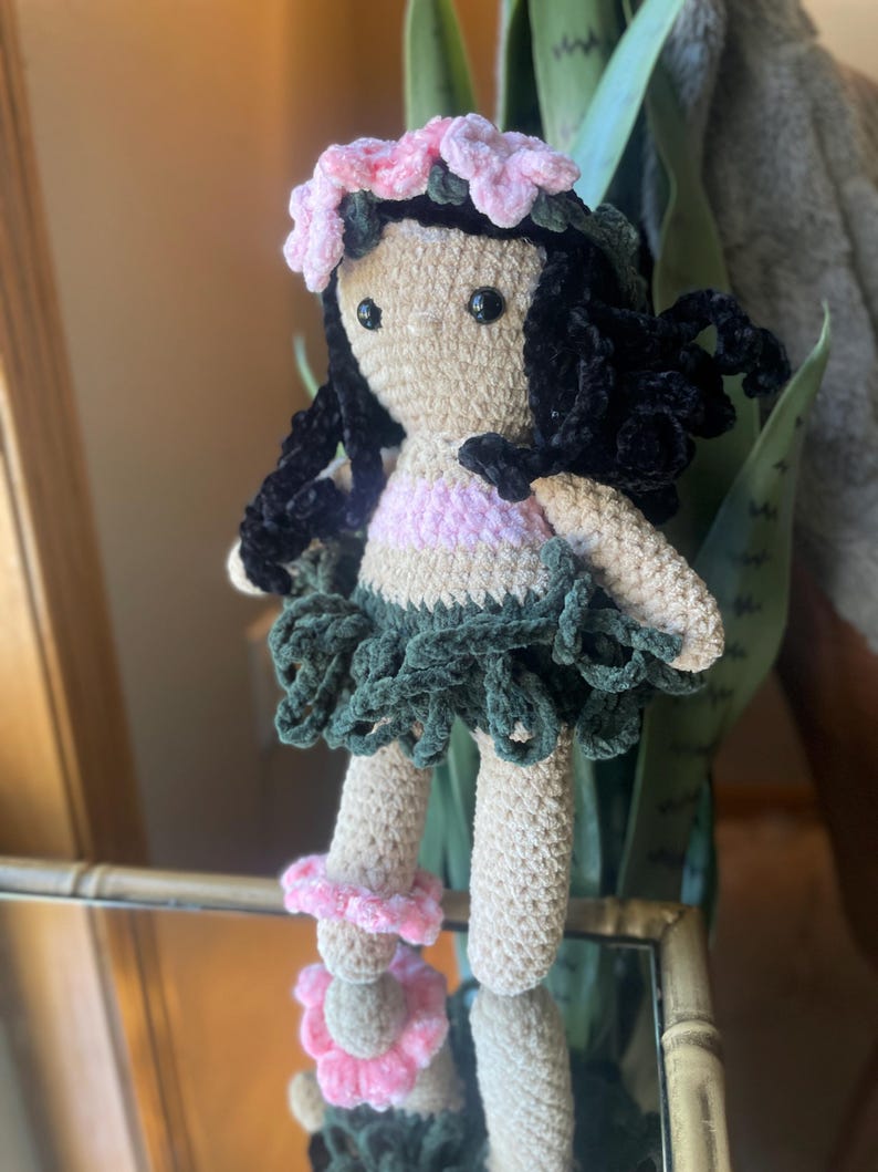 Pode incluir: Boneca de croch&ecirc; com corpo bege, cabelo preto e uma roupa rosa e verde. A boneca tem uma coroa de flores rosa e acess&oacute;rios de tornozelo combinando. A boneca est&aacute; em frente a um espelho.