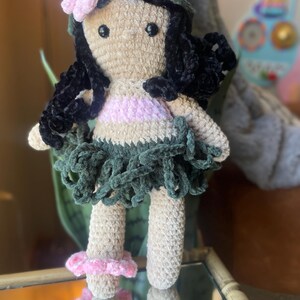 Pode incluir: Boneca de croch&ecirc; feita &agrave; m&atilde;o com corpo bege, cabelo preto e uma roupa rosa e verde. A boneca tem uma coroa de flores rosa e uma saia verde. As pernas da boneca t&ecirc;m detalhes de flores rosa. A boneca &eacute; uma representa&ccedil;&atilde;o de uma dan&ccedil;arina de Hula havaiana.