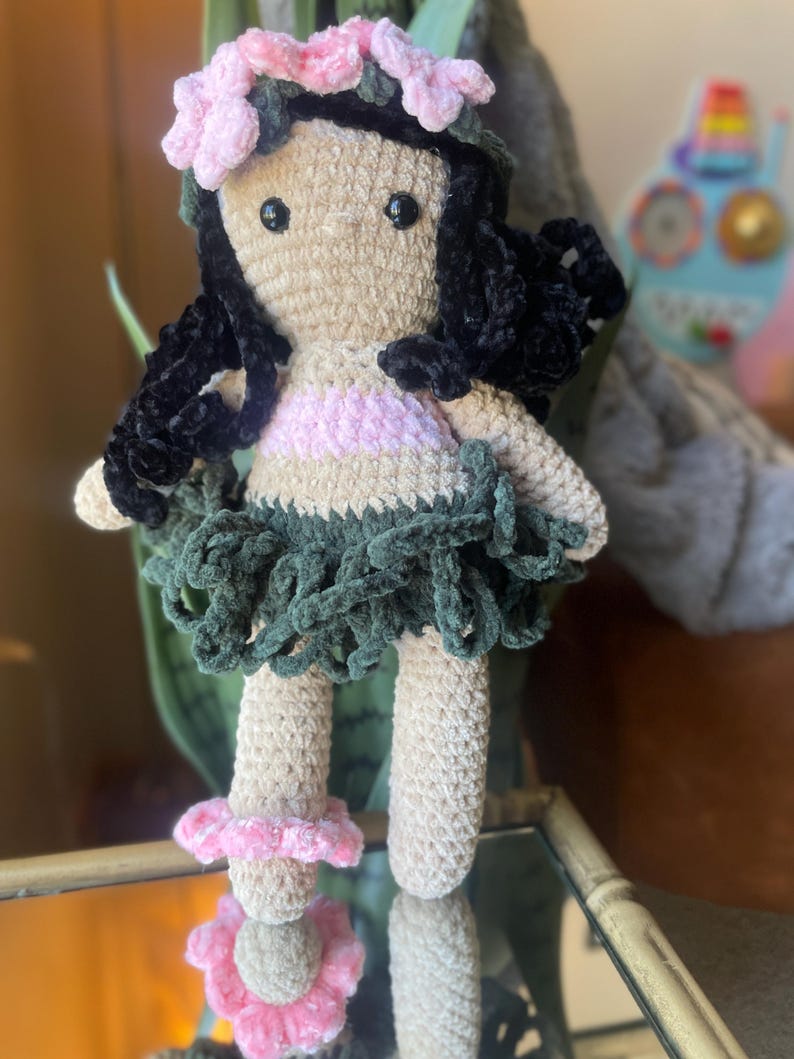 Pode incluir: Boneca de croch&ecirc; feita &agrave; m&atilde;o com corpo bege, cabelo preto e uma roupa rosa e verde. A boneca tem uma coroa de flores rosa e uma saia verde. As pernas da boneca t&ecirc;m detalhes de flores rosa. A boneca &eacute; uma representa&ccedil;&atilde;o de uma dan&ccedil;arina de Hula havaiana.