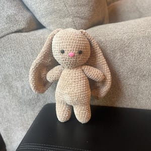 May include: A handmade, beige crochet bunny with long, floppy ears, small arms, and a pink nose. The plush toy is standing on a black surface, with a grey couch in the background. The bunny is a soft, cuddly toy.
