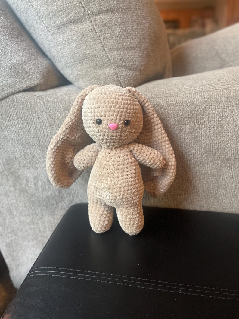 May include: A handmade, beige crochet bunny with long, floppy ears, small arms, and a pink nose. The plush toy is standing on a black surface, with a grey couch in the background. The bunny is a soft, cuddly toy.